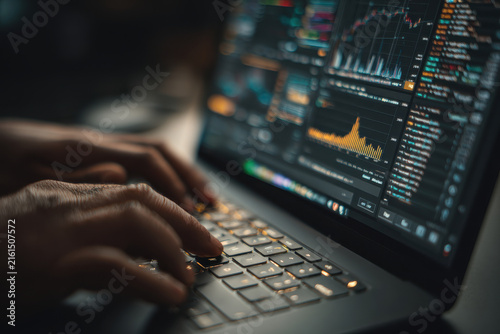 Hands typing code on laptop keyboard, developing trading platform with candlestick charts and data analysis