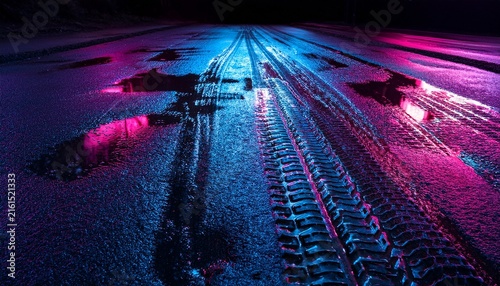 low angle close up of a dark textured asphalt or dirt ground with deep tire tracks and puddles illuminated by vibrant pink and blue neon lights in a moody night scene