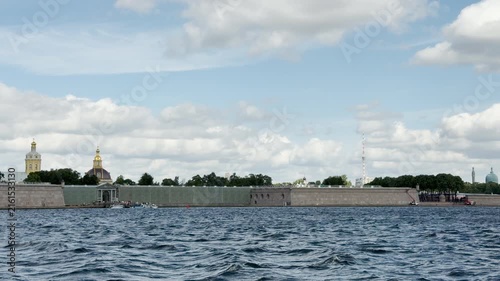 Stone fortress walls stand along the Neva River in Saint Petersburg. Golden domes rise beneath a cloudy blue sky. This magnificent view brings a sense of wonder and calm.