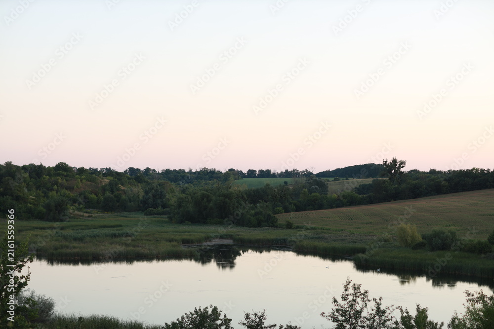 Obraz premium Scenic evening landscape of a calm lake surrounded by rolling hills at early dusk