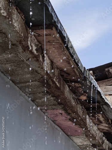 A vertical low-angle macro shot of water dripping from an old rustic roof edge against a blue sky