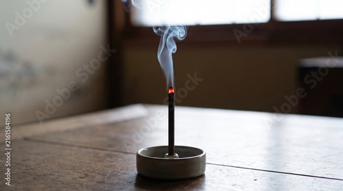 Burning incense stick releasing curling smoke in a ceramic holder on a wooden table. Calm meditation, relaxation, mindfulness, spirituality, and home wellness concept.