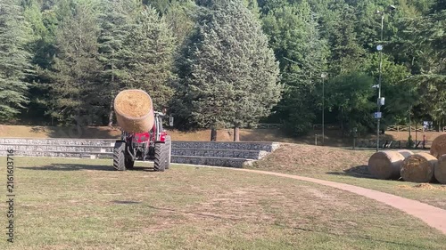 Horizontal slow motion of Tractor loader carries a round hay bale across a grassy park toward stacked bales, showing automated summer farm work and hay preparation.