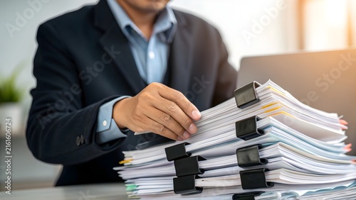 Businessman in a dark suit organizes a massive stack of paper documents with binder clips while working at his office desk.
