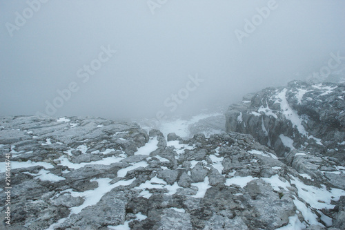 A wide horizontal shot of a rugged limestone mountain plateau covered with patches of snow and shrouded in thick fog in the Dachstein massif of Austria