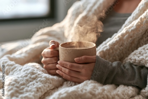 Close up of unrecognizable person wrapping in white knitted blanket. Female hands hold ceramic cup with thick rising steam. Cozy indoor mood for organic herbal tea marketing and winter wellness ads.