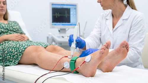 Doctor with blue gloves performs a nerve conduction EMG study on a female patient's leg with electrodes attached