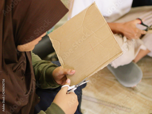 Close up of a young student wearing a hijab cutting a piece of cardboard with scissors. Creative DIY craft, eco-friendly school project, and handmade upcycling concept.