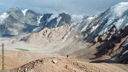 Distant drone view of a woman near Molodezhny Peak above Mynzhylky Plateau, Kazakhstan, with the surrounding mountain range and Tuyuk-Su and Molodezhny glaciers behind her.
