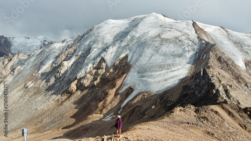 Woman in a cap and shirt standing on Malysh Peak near Almaty, Kazakhstan, in August, with the mountain range and Tuyuk-Su and Molodezhny glaciers in the background.