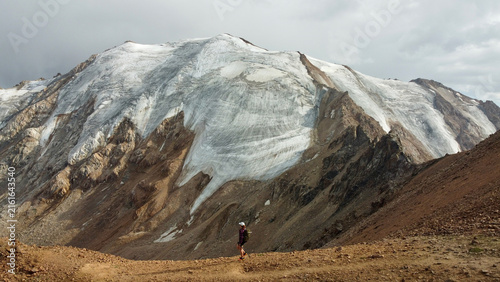 Woman in a cap and shirt hiking along a ridge near Molodezhny Peak by Almaty, Kazakhstan, in August, with the mountain range and Tuyuk-Su and Molodezhny glaciers behind her.