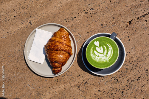 Overhead flat lay of a flaky butter croissant on a ceramic plate beside a matcha green tea latte with leaf-shaped foam art on a stone tabletop.