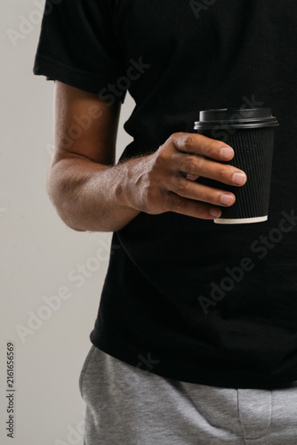 Fotografie A young man with a slender figure, dressed in a black T-shirt, holds an eco-friendly paper cup for hot coffee