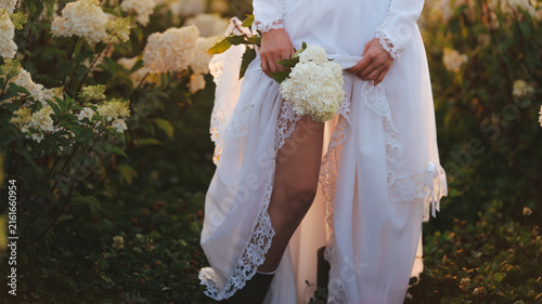 Fotografie Bride in white dress holding bouquet amidst blooming hydrangeas at sunset