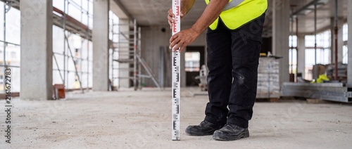 Construction worker holding leveling rod measuring concrete floor elevation inside unfinished building site interior project with worker high visibility