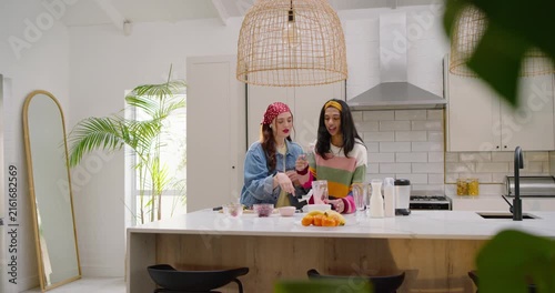 Female friends bandana pointing Indian woman pouring mix into jar at kitchen island making drink