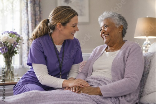 Female nurse in purple scrubs talking to a smiling senior African American woman lying in bed at home.