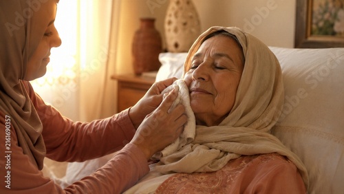 Young woman caregiver wiping the face of an elderly woman in a hijab with a soft towel.