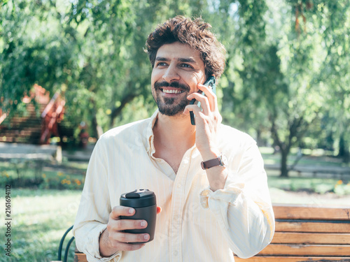 Adult man sitting on a park bench with a takeaway coffee while looking at his smartphone. The sunny outdoor setting represents mobile technology, digital communication, relaxation, social media and mo