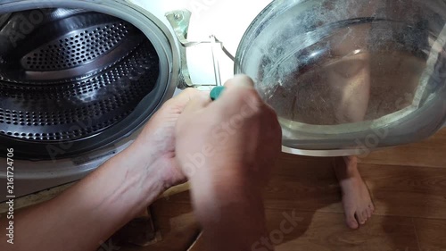 Person repairing washing machine drum at home, close-up of hands removing black perforated tub with transparent door component.