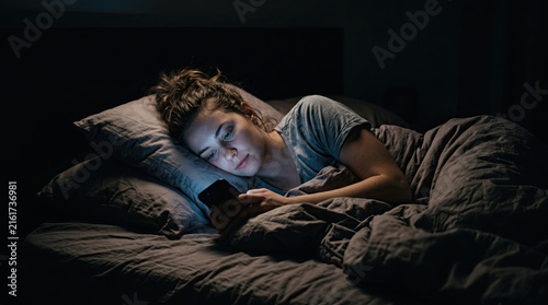 Woman lying in bed using smartphone at night under the covers during sleepless night	