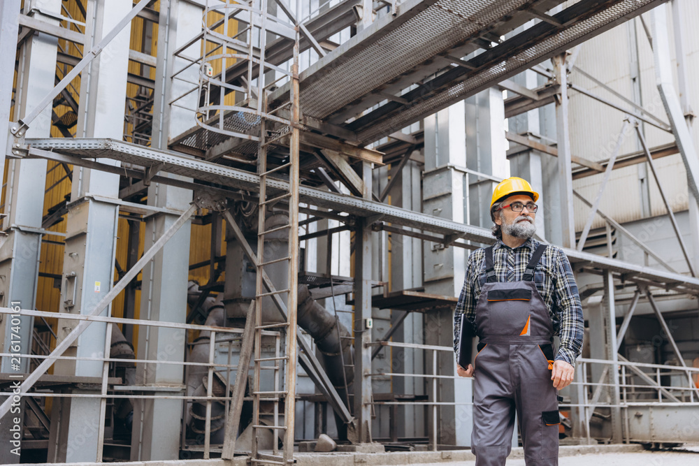 Fototapeta premium Male worker wearing hard hat and uniform, carrying a tablet, supervising operations at a large grain processing plant