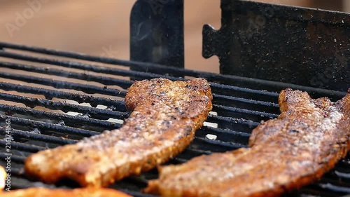 Close-up of seasoned pork belly slices sizzling on hot BBQ grill outdoors with smoke and flames
