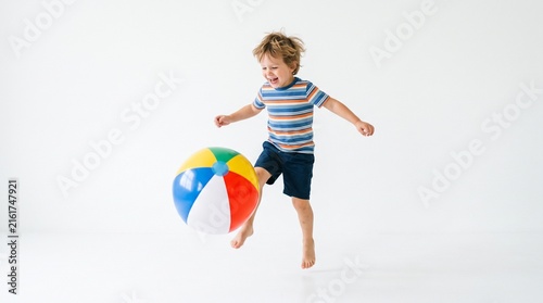 Joyful young boy in striped shirt and shorts kicking a colorful beach ball while jumping in a studio