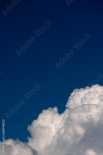 Deep blue vertical sky with fluffy white cumulus cloud in bottom corner. Minimalist cloudscape featuring negative space, bright sunlight, natural weather background, clear air, and summer day scenery.