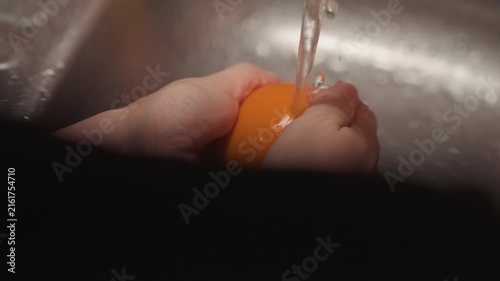 Close up of hands washing orange under running water in sink. Fruit is rotated and rinsed thoroughly with visible wet skin. Metallic kitchen setting provides context