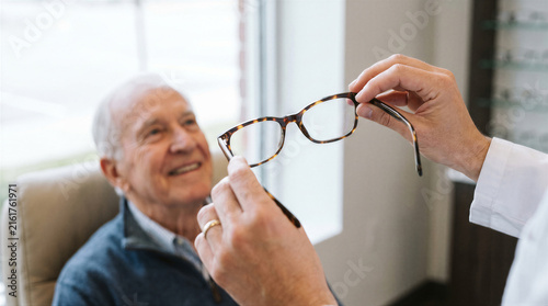 Optometrist holding up a pair of tortoise shell eyeglasses for an elderly male patient during a vision consultation