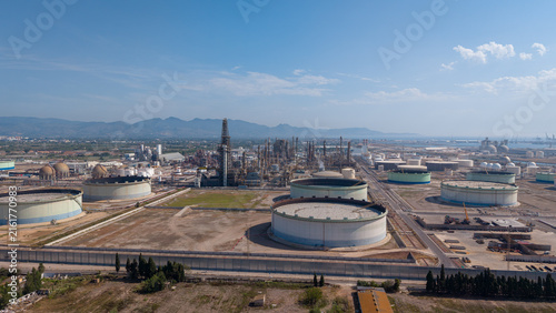 Expansive oil refinery complex with large cylindrical storage tanks, distillation towers, and industrial infrastructure set against mountains and blue sky