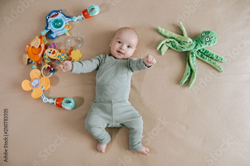 Cute 3 month baby boy lying among various playful colorful toys on beige blanket. Infant surrounded by vivid objects participating actively in sensory exploration and learning. 
