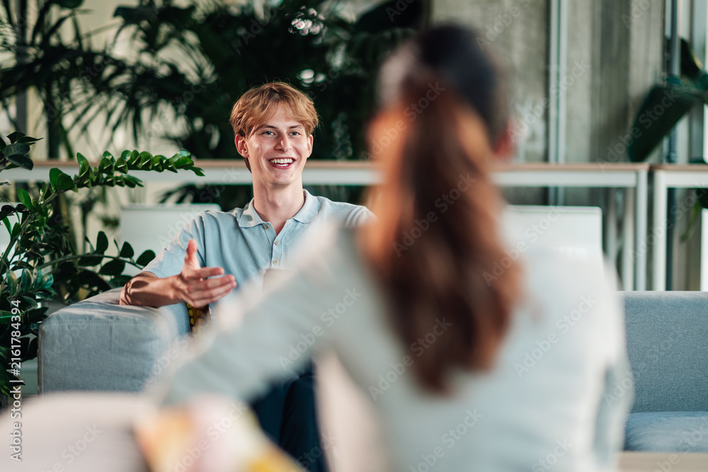 Fototapeta premium Young man smiling and gesturing while talking with a colleague in an office lounge, collaborating in a modern coworking space