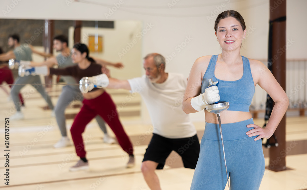 Fototapeta premium Young woman is engaged in fencing in a modern sports club. Woman with a rapier in the training hall prepares for a lesson while other athletes practice the technique