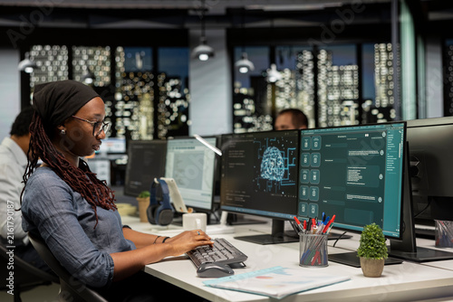 Startup worker uses a large language model application for coding help while an African American woman interacts with AI LLM chatbot software on a PC in a software development workplace.