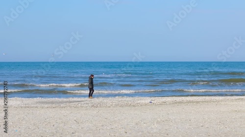 Lone woman walking alone on a deserted beach. Solitary woman in a warm jacket taking a peaceful walk along the seashore on a cold, sunny day. The calm blue sea create a melancholic atmosphere