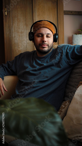 Young Latino man sitting on his sofa with his eyes closed and wearing headphones, listening to music and enjoying a peaceful, relaxing moment at home.