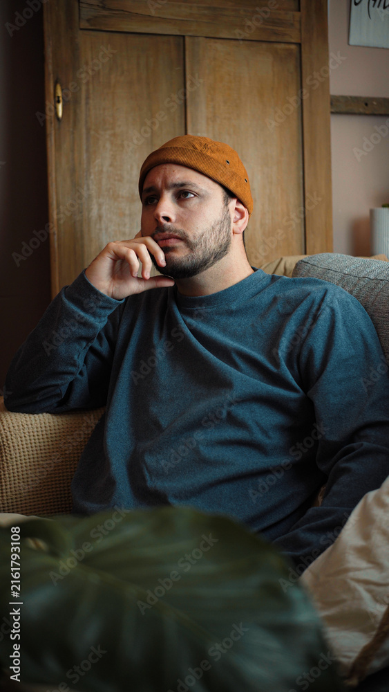 Obraz premium Young Latino man sitting on his sofa, looking very thoughtful and lost in contemplation during a quiet moment at home.
