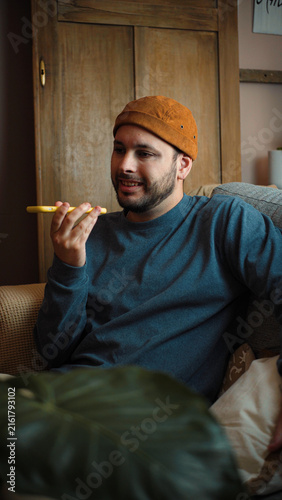 Young Latino man sitting on his sofa and recording or sending a voice message through his smartphone, communicating from the comfort of his home.