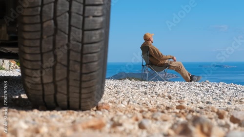 Man relaxing on camping chair enjoying seascape view. Adventurous traveler sitting in a foldable chair next to his off-road vehicle, relaxing and contemplating the stunning seascape