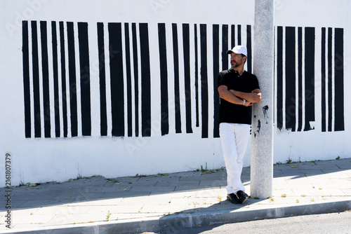 Obraz na płótnie A tall, stylish, dark-haired, thin, 40-year-old white man with a beard is wearing white pants, black shoes, a black polo shirt