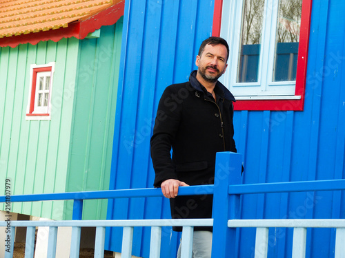 Canvastavla White man, dark-skinned, thin, 40 years old, casually dressed, posing against a backdrop of colorful wooden houses