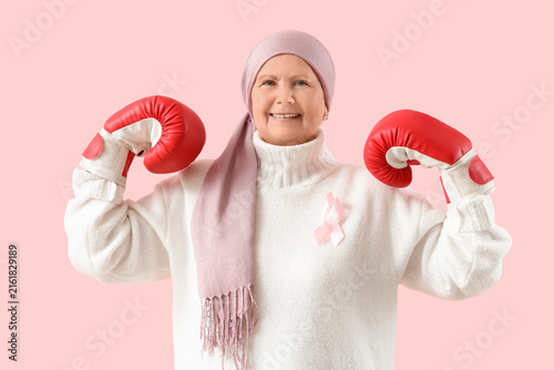 Senior woman after chemotherapy in boxing gloves with pink ribbon on color ba...