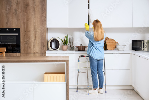 Mature woman with walker and pp-duster cleaning cupboard in kitchen, back view