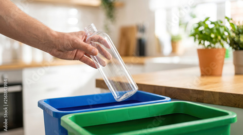 Hand places a clear bottle into a blue recycling bin next to a green bin in a bright kitchen
