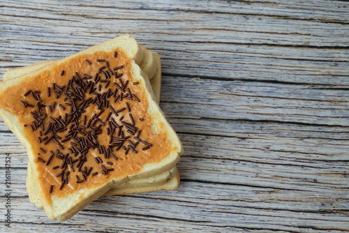 White breads slice topped peanut butter with dark sprinkles rests beside plain bread. Wooden surface shows grain, rustic texture, natural tones. Simple, clean composition highlights food's 