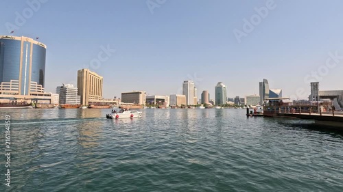 A boat sails across a wide bay in Dubai during daytime, with modern skyscrapers forming the backdrop. The scene captures the urban waterfront and calm water.