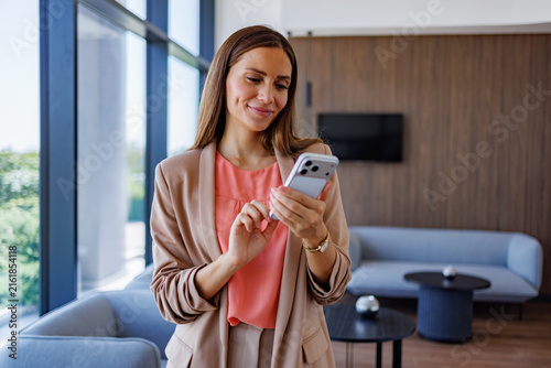Businesswoman standing in modern office lobby, smiling and typing on smartphone, connecting and communicating