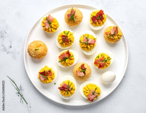 White platter of deviled eggs, some topped with bacon and chives, others breaded